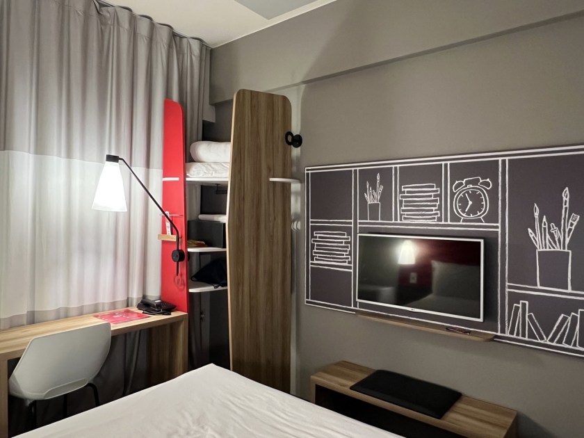 À esquerda, mesa de escritório com abajur. No centro, pequeno quarda-roupas aberto. E à direita, televisão na parede com painel preto e ilustrações em giz atrás.