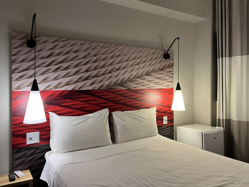 Quarto de hotel com lençois brancos na cama, cabeceira com impressão de cordas vermelhas e brancas e abajures pendentes com luzes acesas.