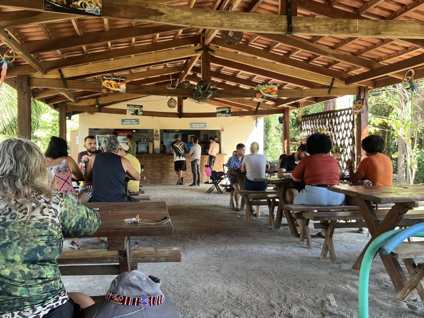 Restaurante com cobertura de telhado e diversas pessoas sentadas em bancos e mesas de madeira.