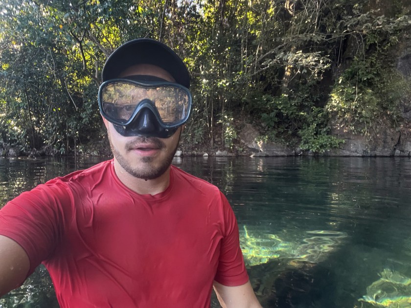 Homem dentro de uma lagoa com mata atrás, usando boné, máscara de snorkel e camisa vermelha.
