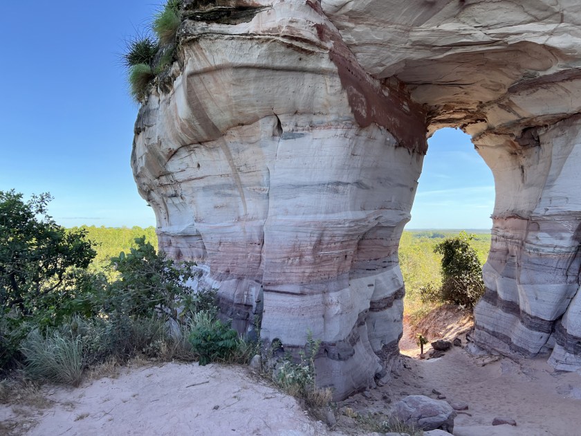 Formação rochosa com buraco com vista para a vegetação do cerrado e céu azul ao fundo.