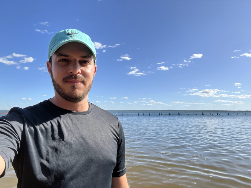 Foto de homem com camisa preta e boné verde em lago e céu azul.