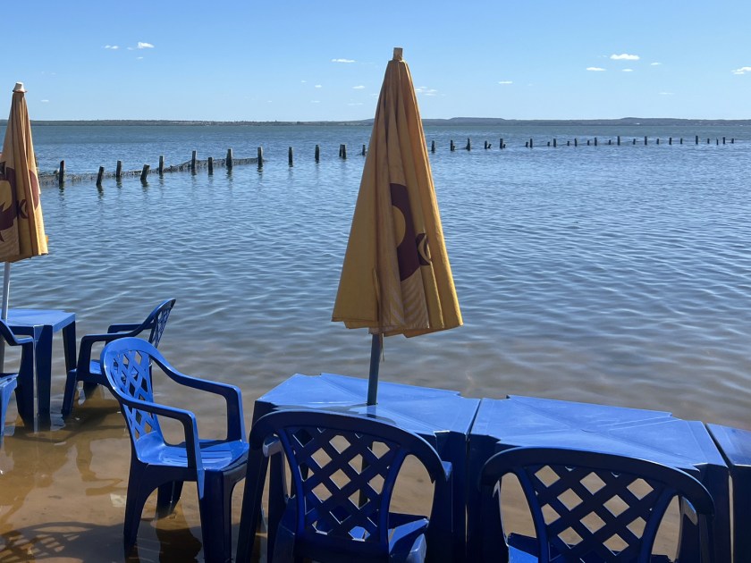 Mesas e cadeiras de plástico azul e guarda-sol amarelo posicionados nas águas mansas e rasas de um lago com céu limpo ao fundo.